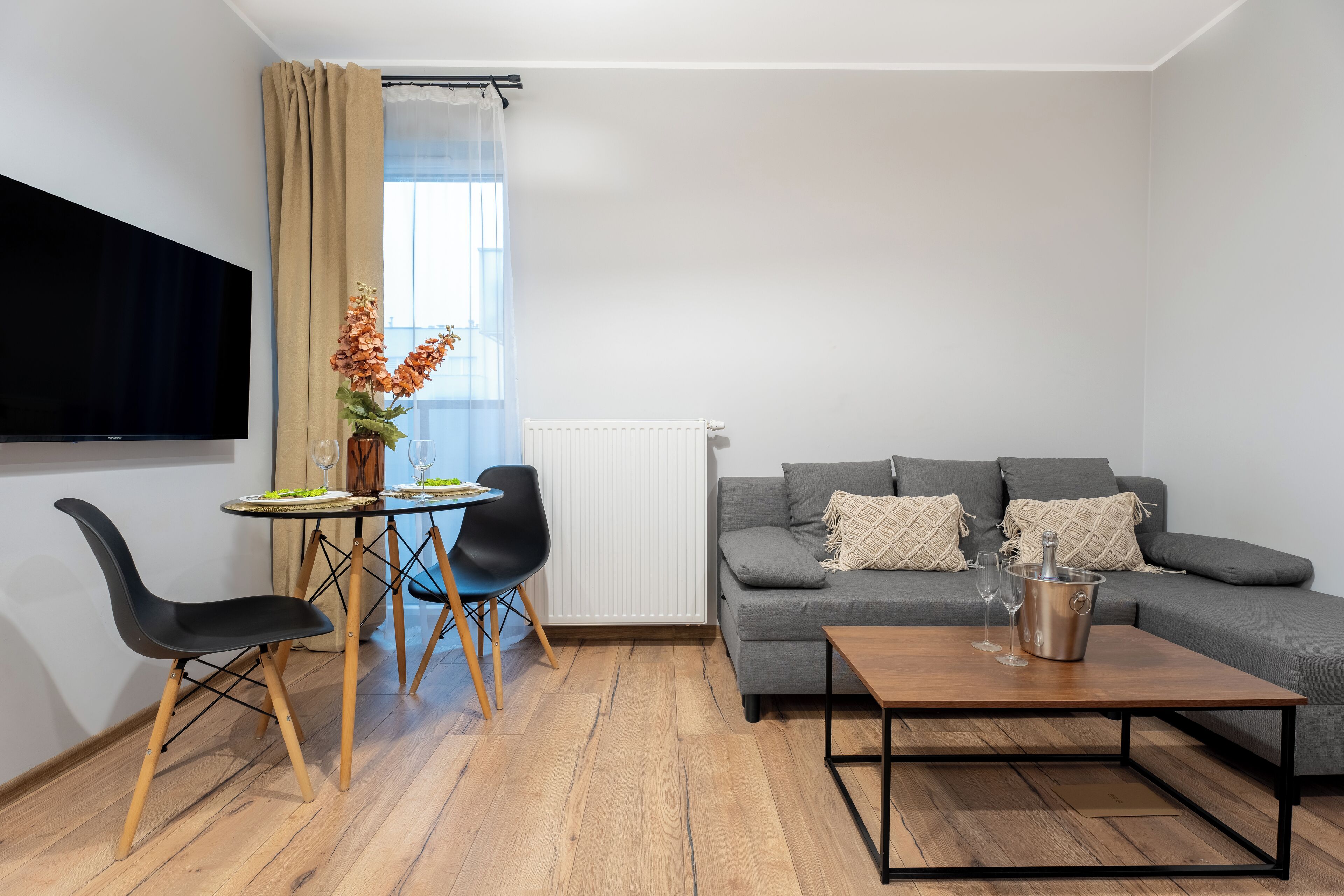 Una sala de estar moderna con un sofá gris, una mesa de centro de madera, un televisor montado en la pared y una zona de comedor con una mesa pequeña y sillas.