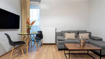 Una sala de estar moderna con un sofá gris, una mesa de centro de madera, un televisor montado en la pared y una zona de comedor con una mesa pequeña y sillas.