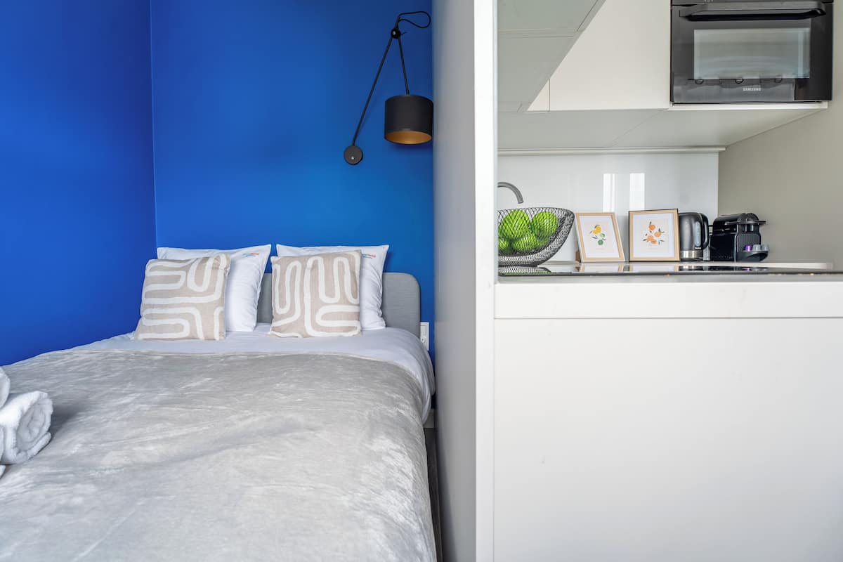 A modern hotel room with a bed, a blue accent wall, a kitchen area with a coffee maker, and a hanging lamp.