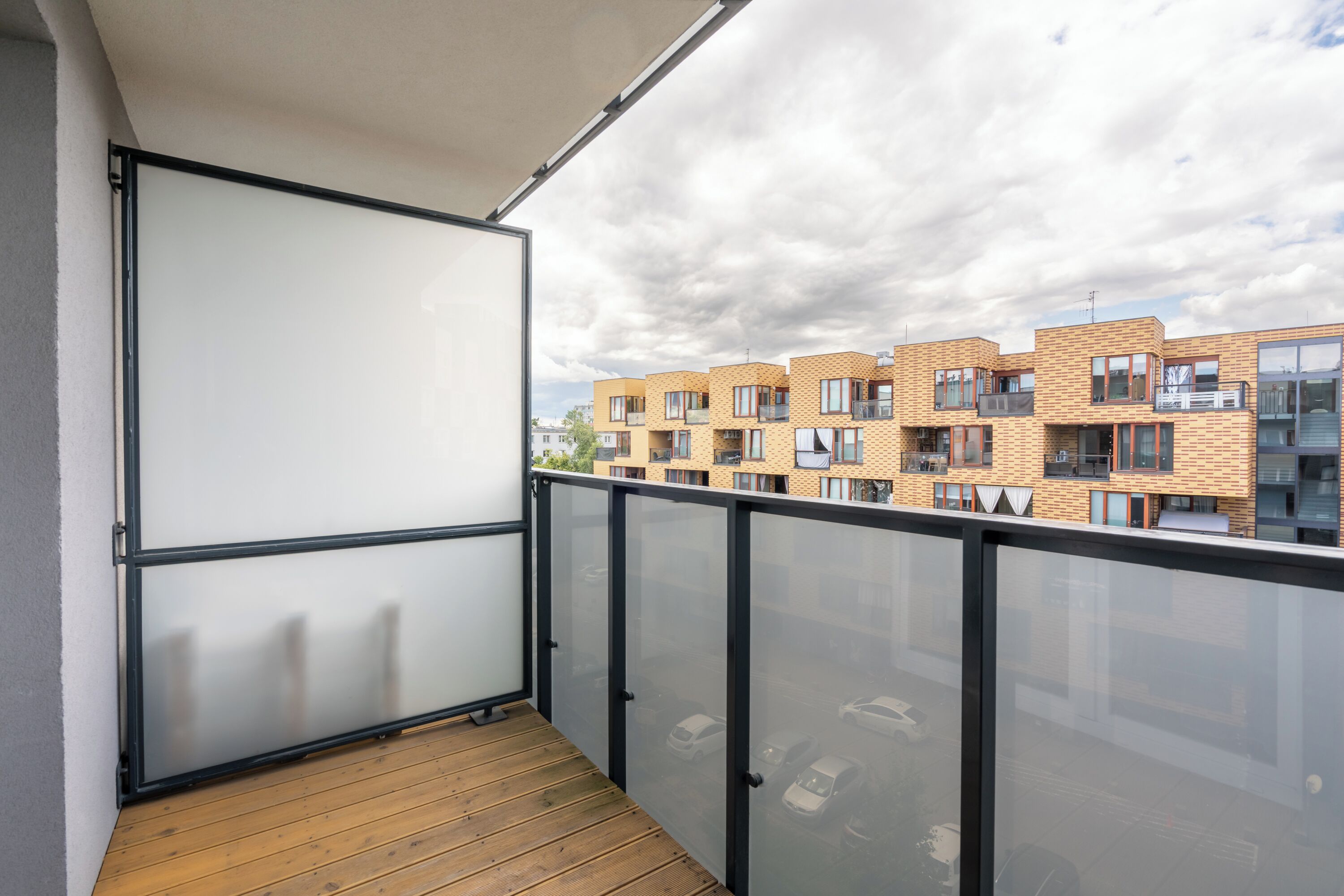 Un balcon doté d’un plancher en bois et d’une rambarde en verre, offrant une vue sur une rangée d’immeubles d’appartements modernes en briques.