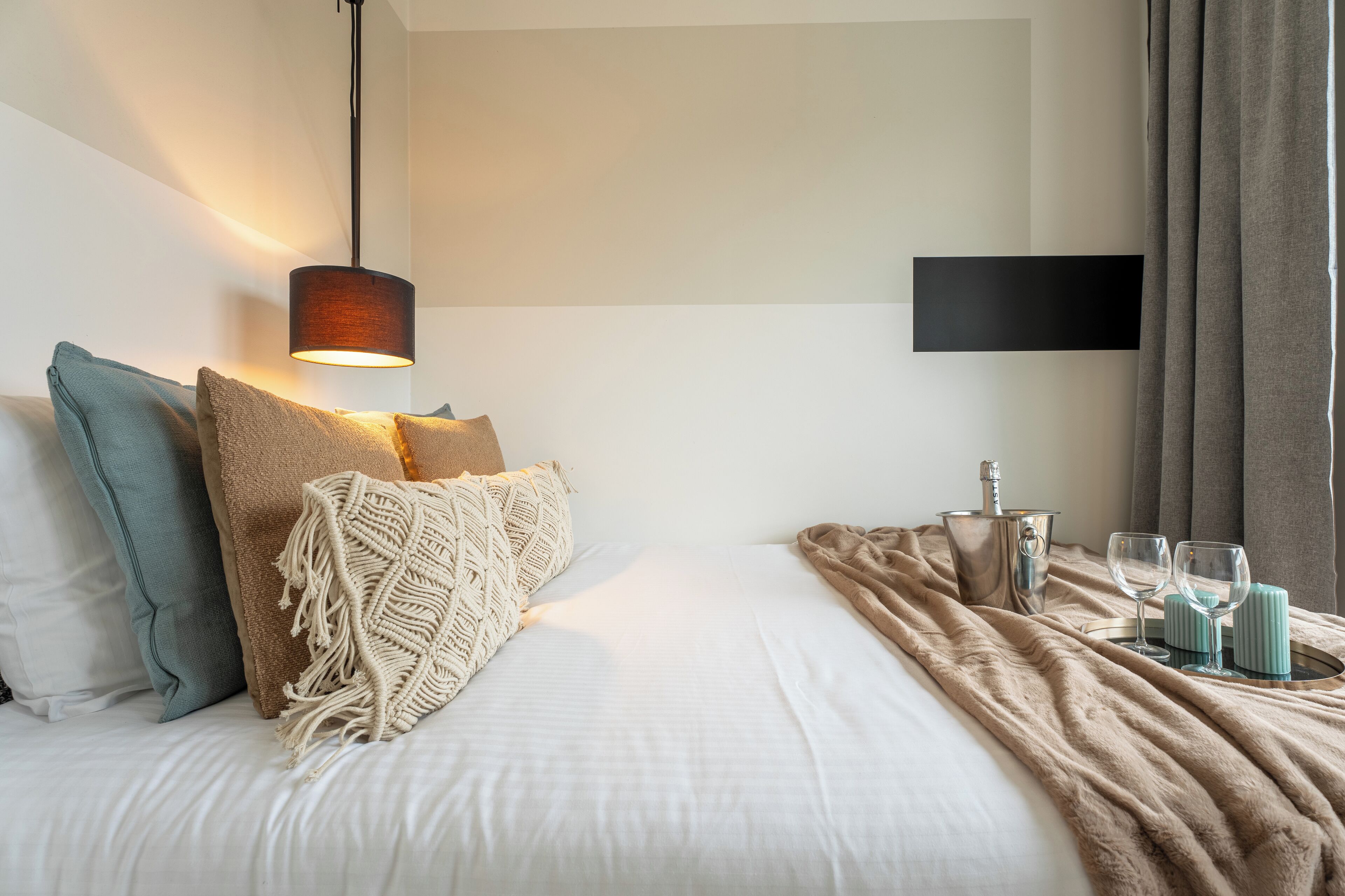 Un lit bien fait, avec une couette blanche, un plaid beige et un couvre-lit en macramé. On y trouve une table de chevet avec une bouteille, deux verres et un plateau. Un téléviseur à écran plat est fixé au mur.