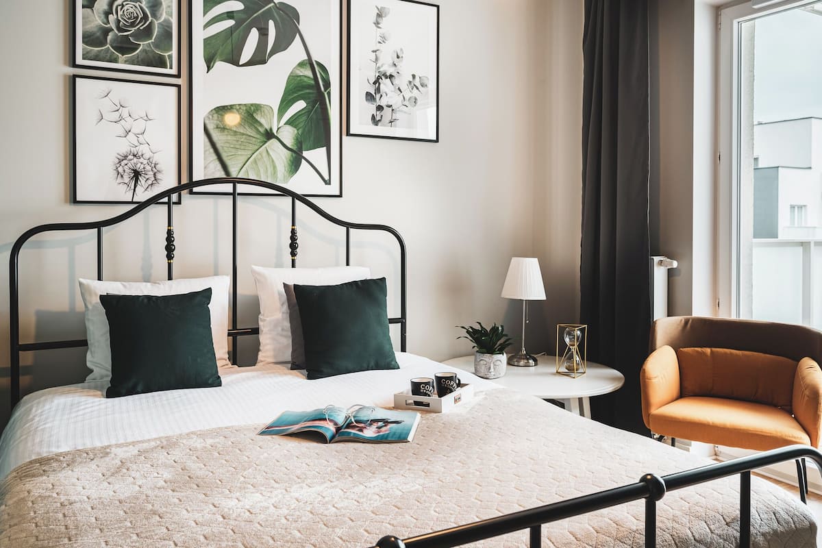 A neatly made bed with a quilt, two pillows, a bedside table with a lamp and a plant, and a chair.