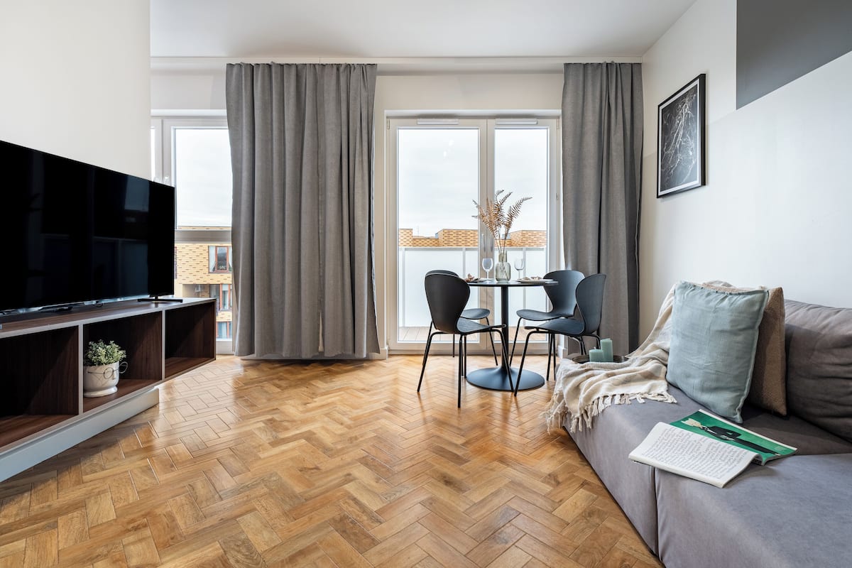 A modern living room with a flat-screen TV, a sofa, a dining table with chairs, and a balcony with curtains.