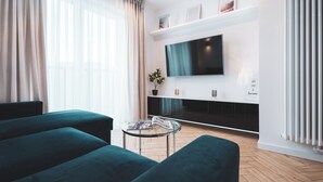 A modern living room with a flat-screen TV mounted on the wall, a glass coffee table, and two green sofas. - Pure Rental Premium Inżynierska (Wrocław)