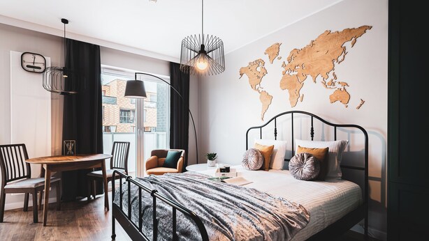 A modern bedroom with a bed, a desk, and a chair. There is a world map on the wall and a window with curtains. - Pure Rental Premium Inżynierska (Wrocław)