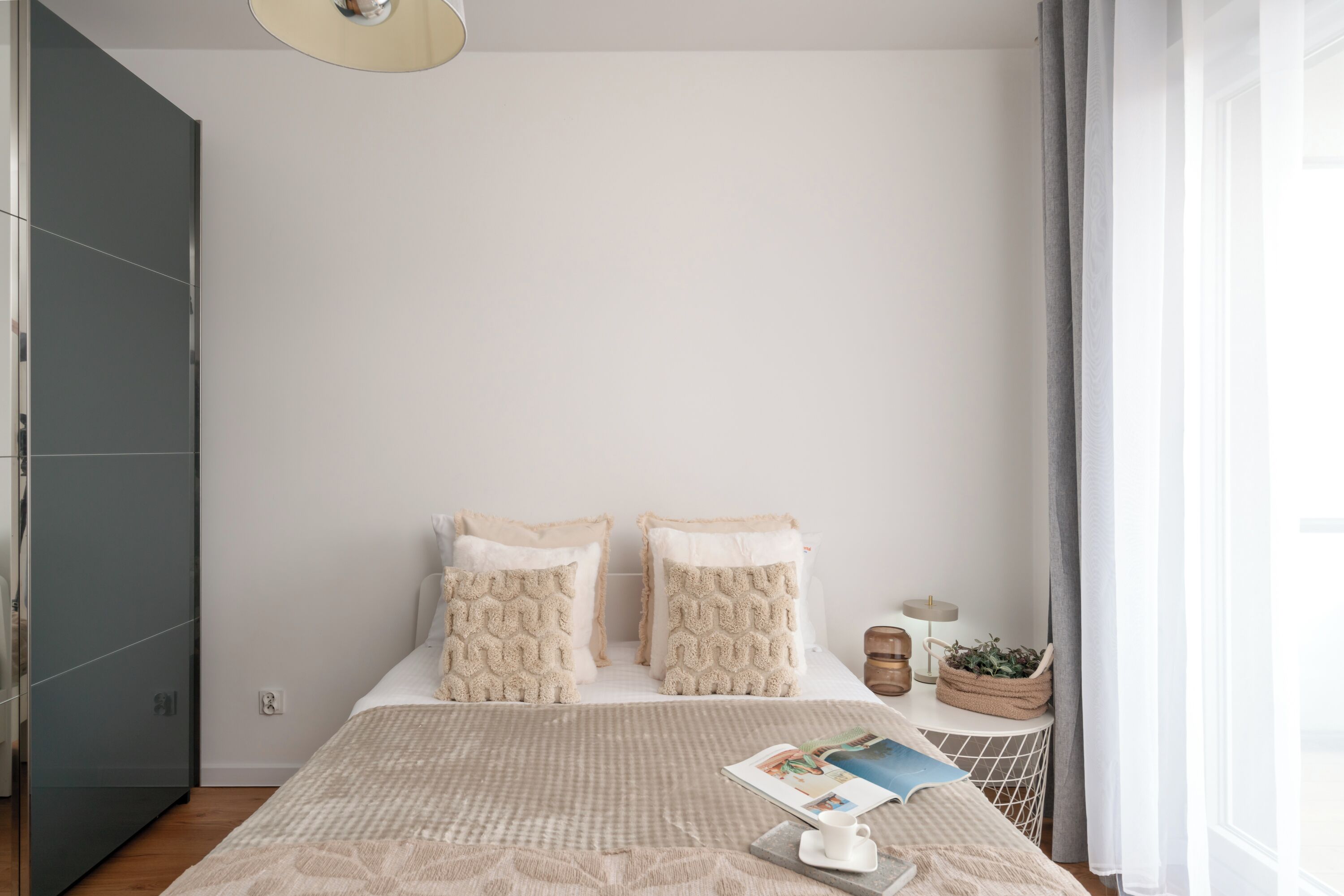 Un dormitorio con cama, almohadas, una mesita de noche con lámpara, una revista y una planta.