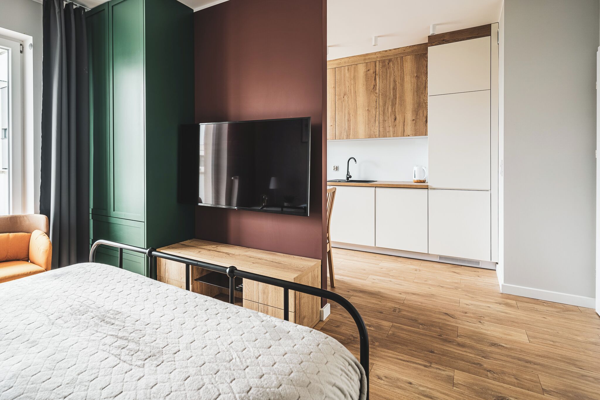 A modern hotel room with a bed, a desk, a television, and a kitchenette.