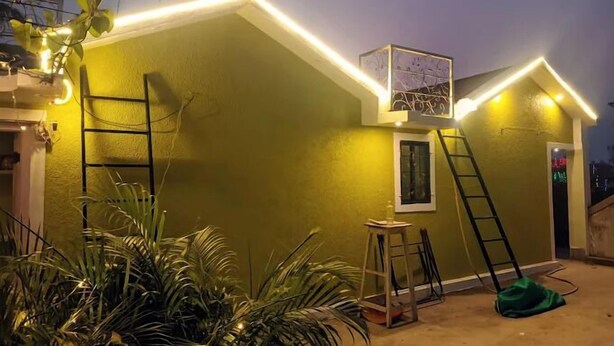 A small, single-story building with a green exterior and a small porch area. There are two ladders leaning against the building, a green hose on the ground, and a small table with a chair. - Pravuprasad Homestay (Bhubaneshwar)