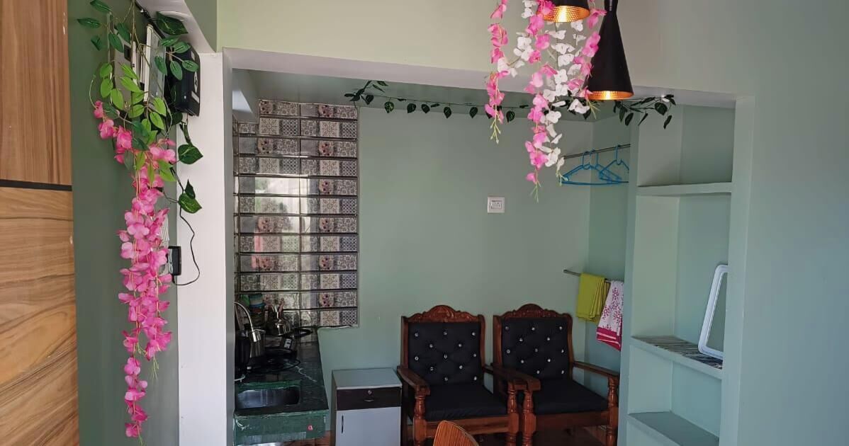 A compact kitchen with a gas stove, a small refrigerator, and a sink. There are two wooden chairs with black cushions and a dining table.