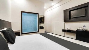A hotel room with a large bed, a TV mounted on the wall, a small desk with a mirror, and a window with a view of the sky. - Hotel OPM & Restaurant (Agra)