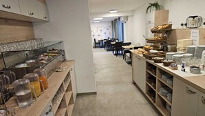 Disposição de buffet com diversos alimentos e bebidas, área de estar com mesas e cadeiras, e balcão com pilhas de pratos e copos.