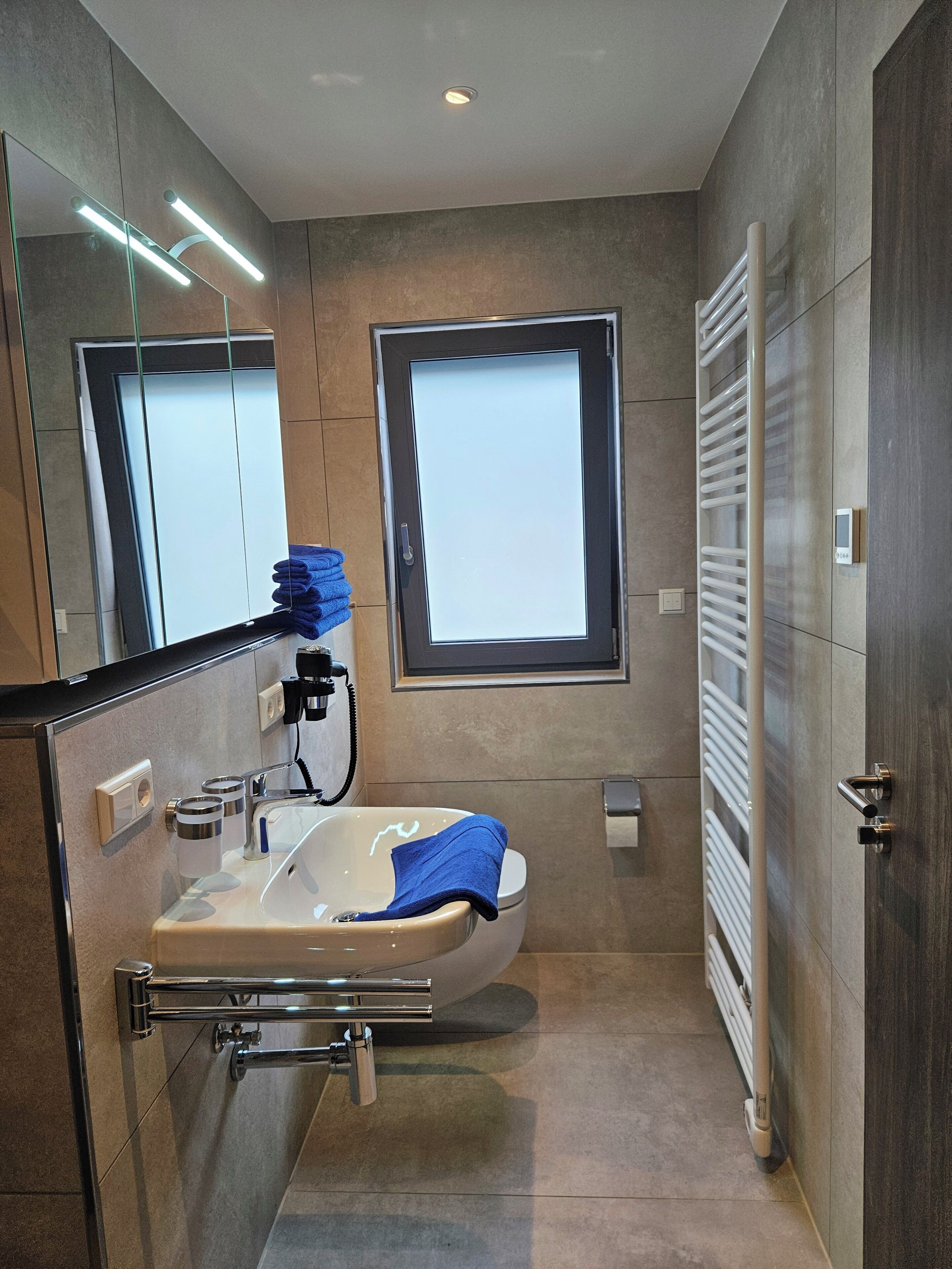 Un baño moderno con un espejo grande, un lavamanos con una toalla, una ventana y un perchero para toallas.