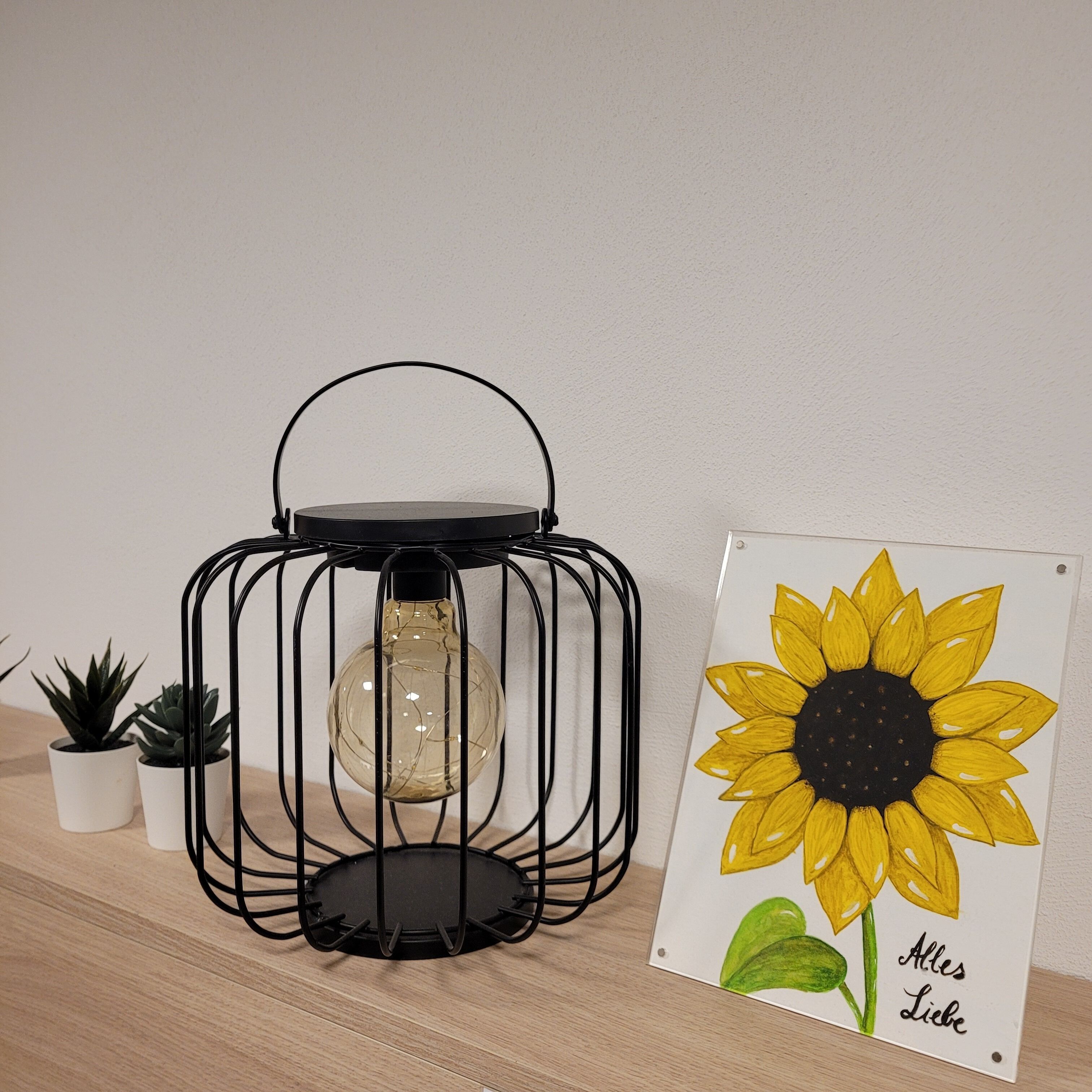 Une lanterne en métal noir avec un globe en verre transparent, deux plantes en pot et une œuvre d’art représentant un tournesol, encadrée et accompagnée d’un texte.