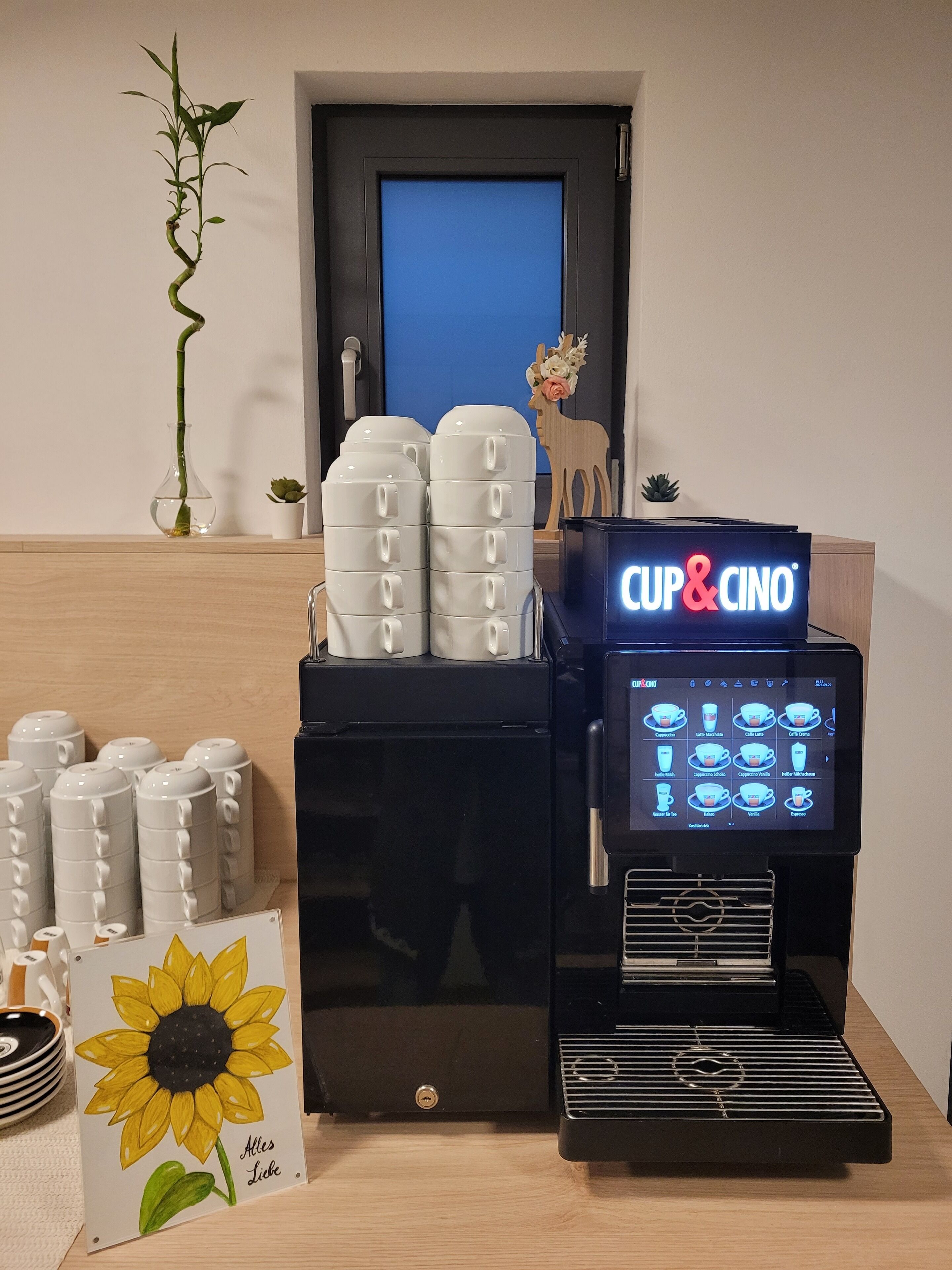 Una cafetera con pantalla digital, pilas de tazas blancas y una obra de arte con girasoles sobre una mesa.