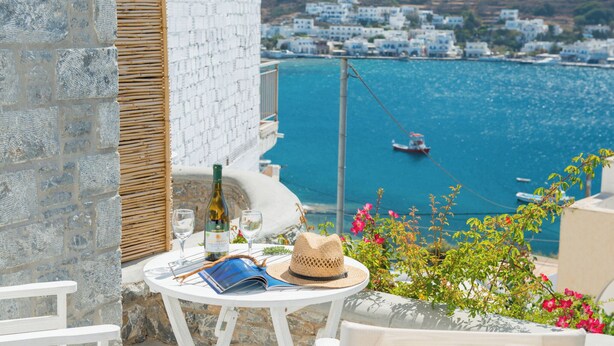 Una terrazza vista mare con un tavolo apparecchiato per due, una bottiglia di vino, un bicchiere da vino, un cappello e un libro.