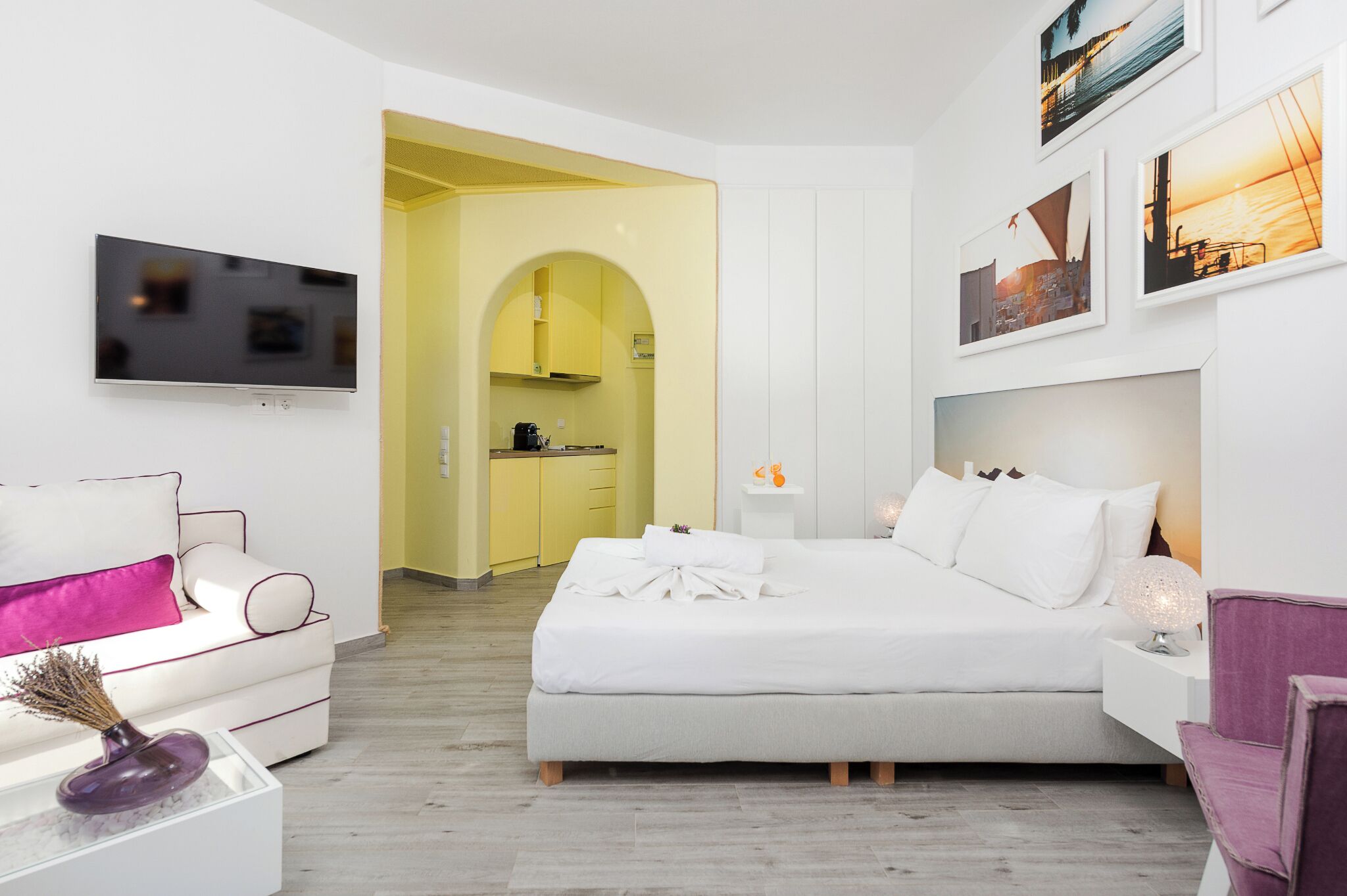 Una habitación de hotel moderna con cama, sofá, televisor y zona de cocina visible a través de un arco.