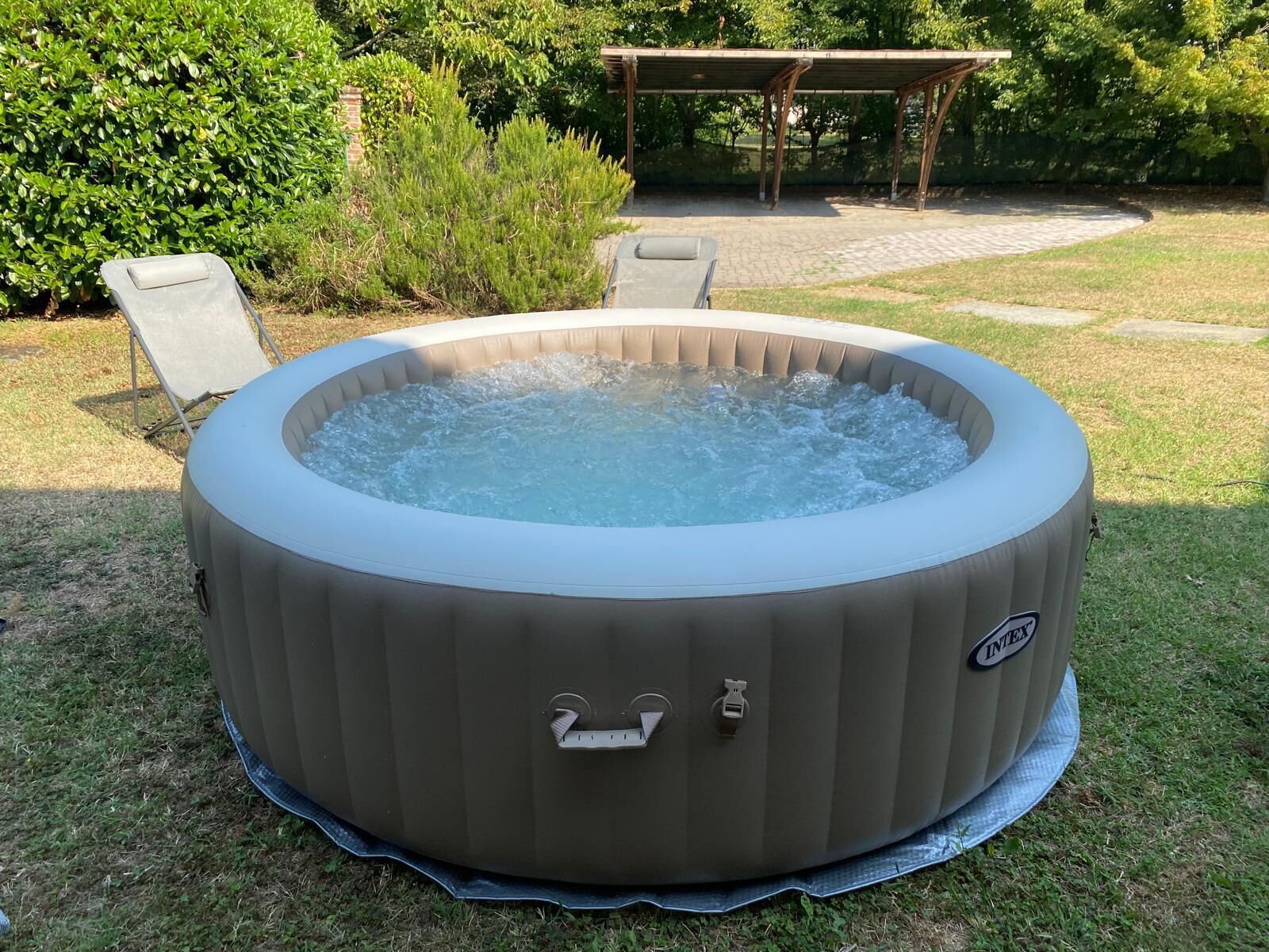 A hot tub with a handle and a label, surrounded by grass and a patio area with chairs.
