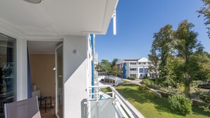 A balcony with a view of a well-maintained lawn, a building with a blue and white facade, and a clear blue sky. - Vacation Rental: “Apartment on the Zingst Pier with a South-Facing Balcony” (Zingst)