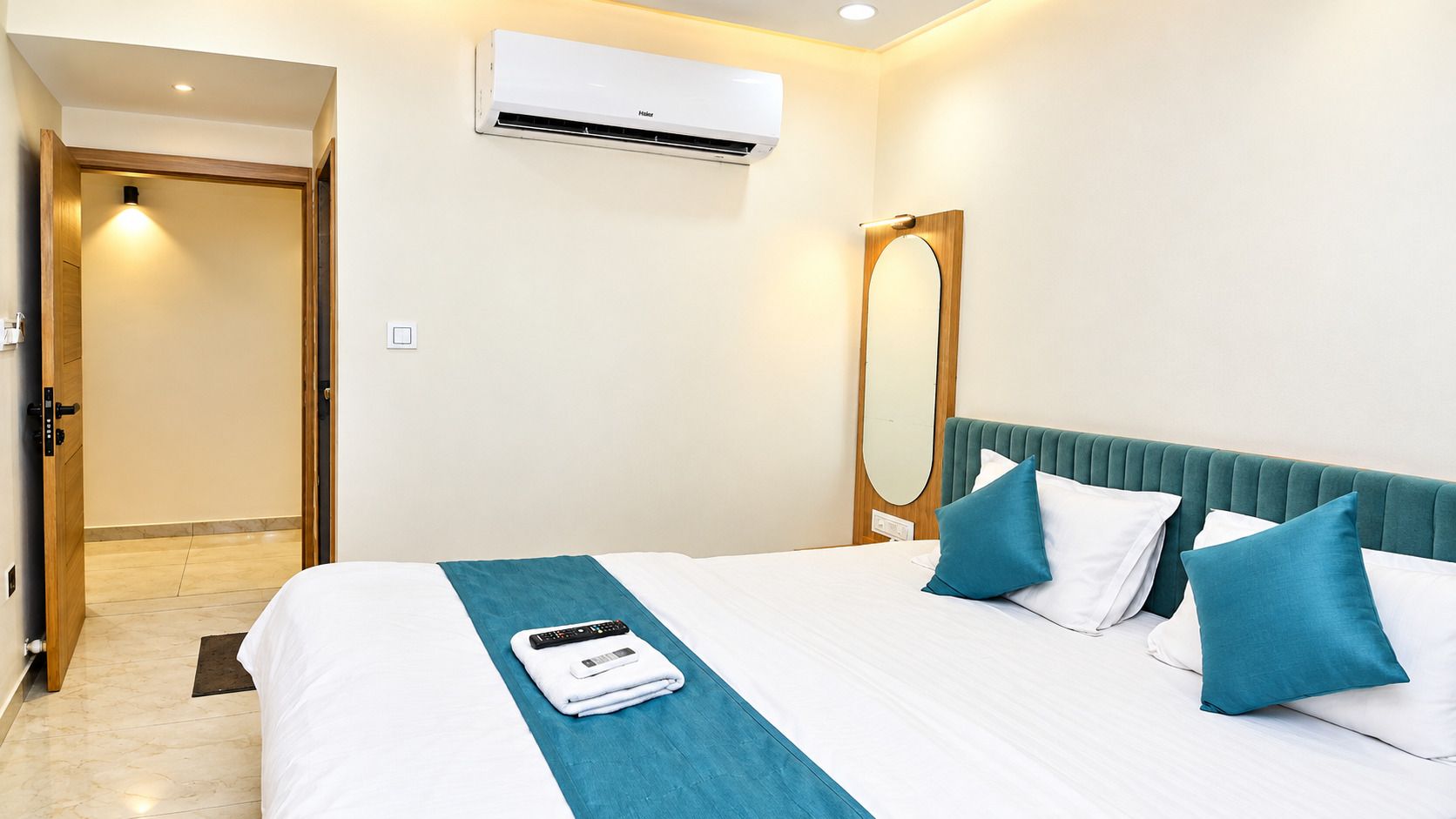 A hotel room with a bed, a remote control, a folded towel, a mirror, and an air conditioning unit.
