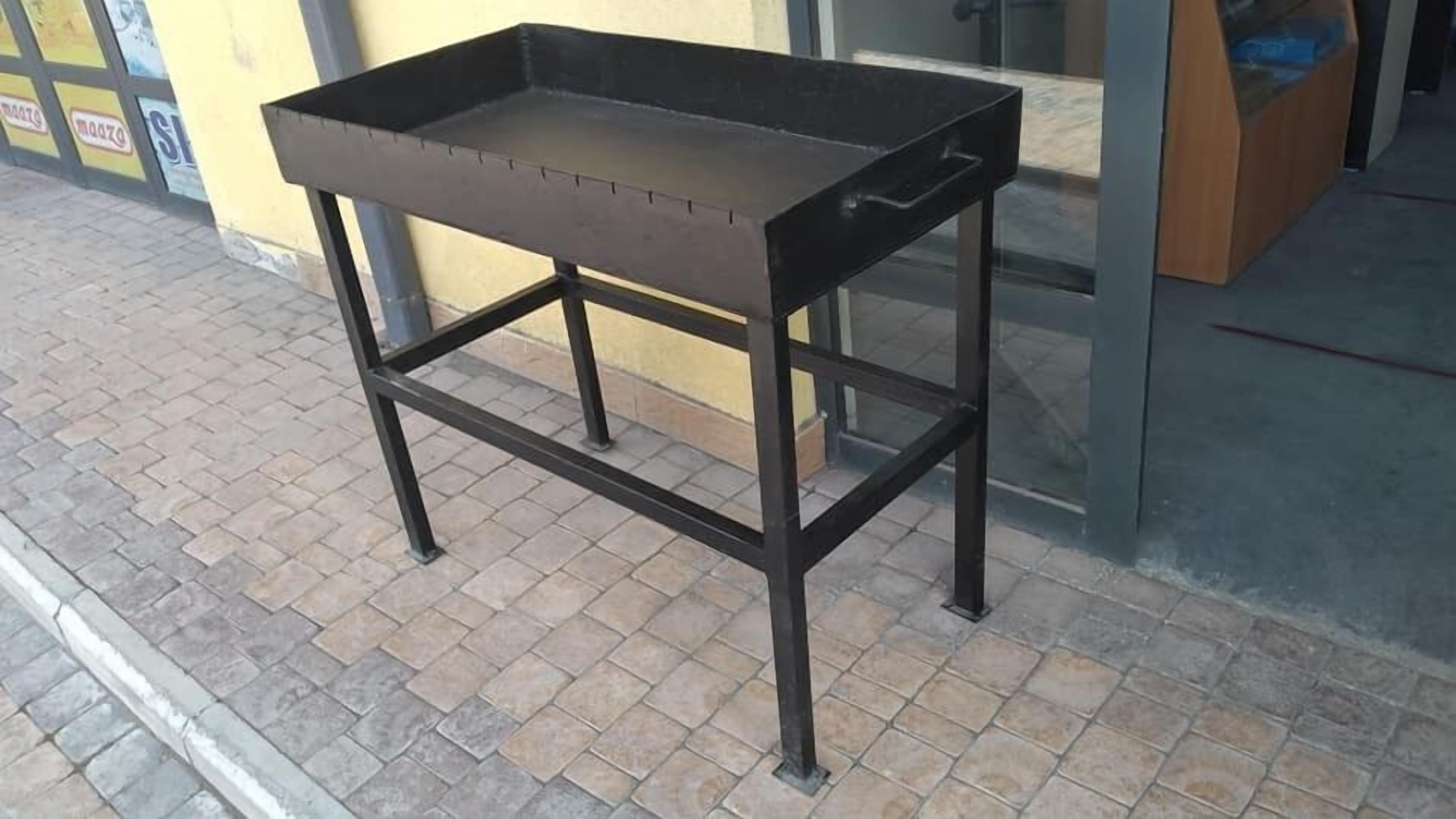 Een metalen tafel met een rechthoekig tafelblad en vier poten, op een betegelde stoep voor een gebouw.