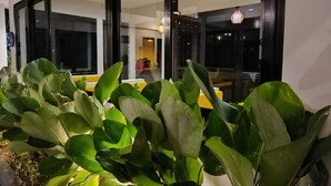 A well-lit indoor area with large windows, a dining area with yellow chairs, and lush green plants in the foreground. - Padinis Villa & Coffee (Sukawati)