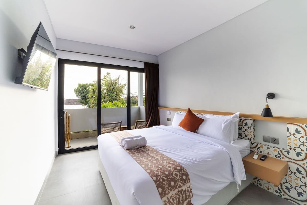 A hotel room with a bed, a bedside table, a TV mounted on the wall, and a sliding glass door leading to an outdoor area.