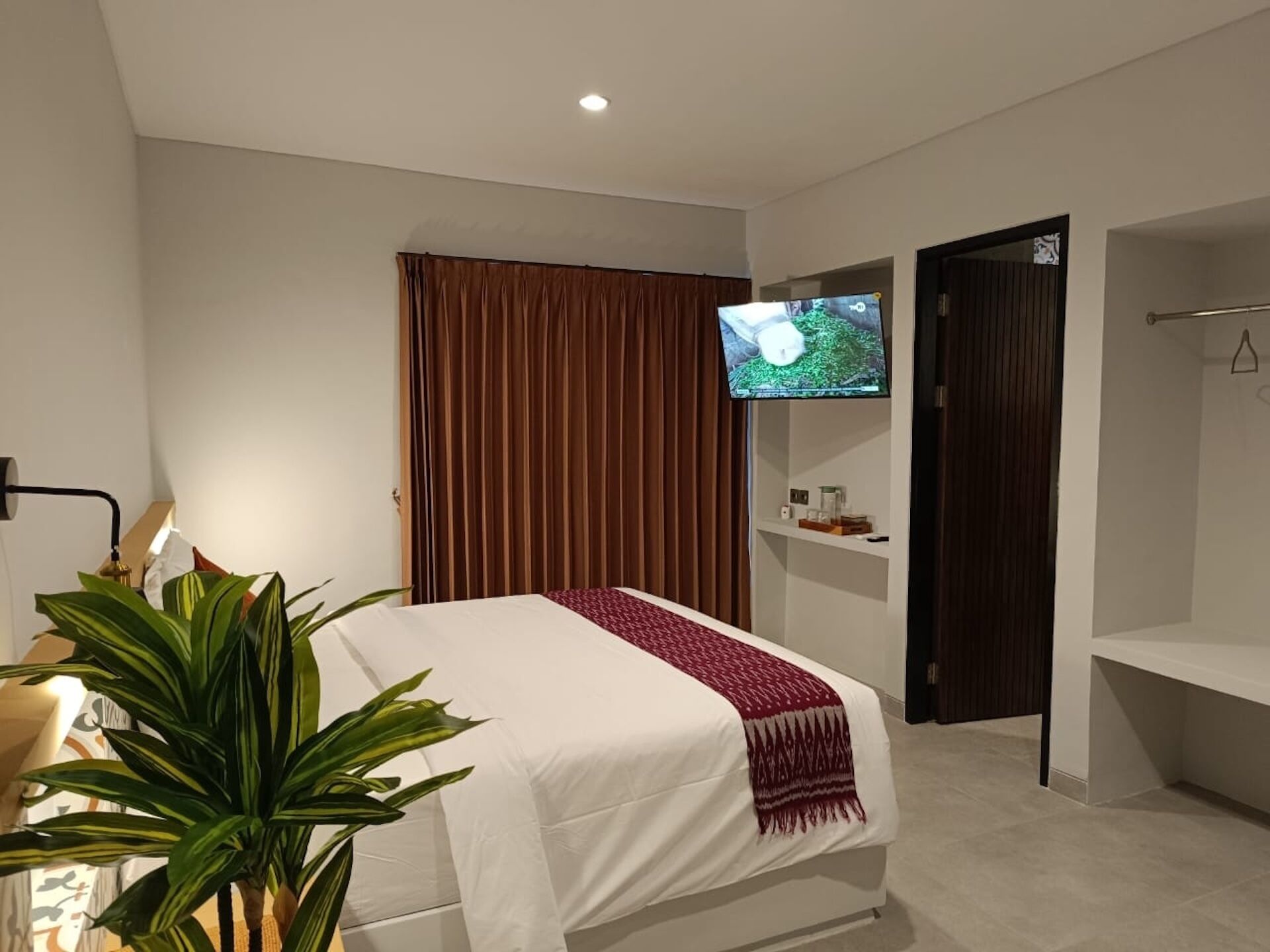 Een moderne hotelkamer met een bed, een aan de muur bevestigde televisie, een plant en een deur naar een andere kamer.