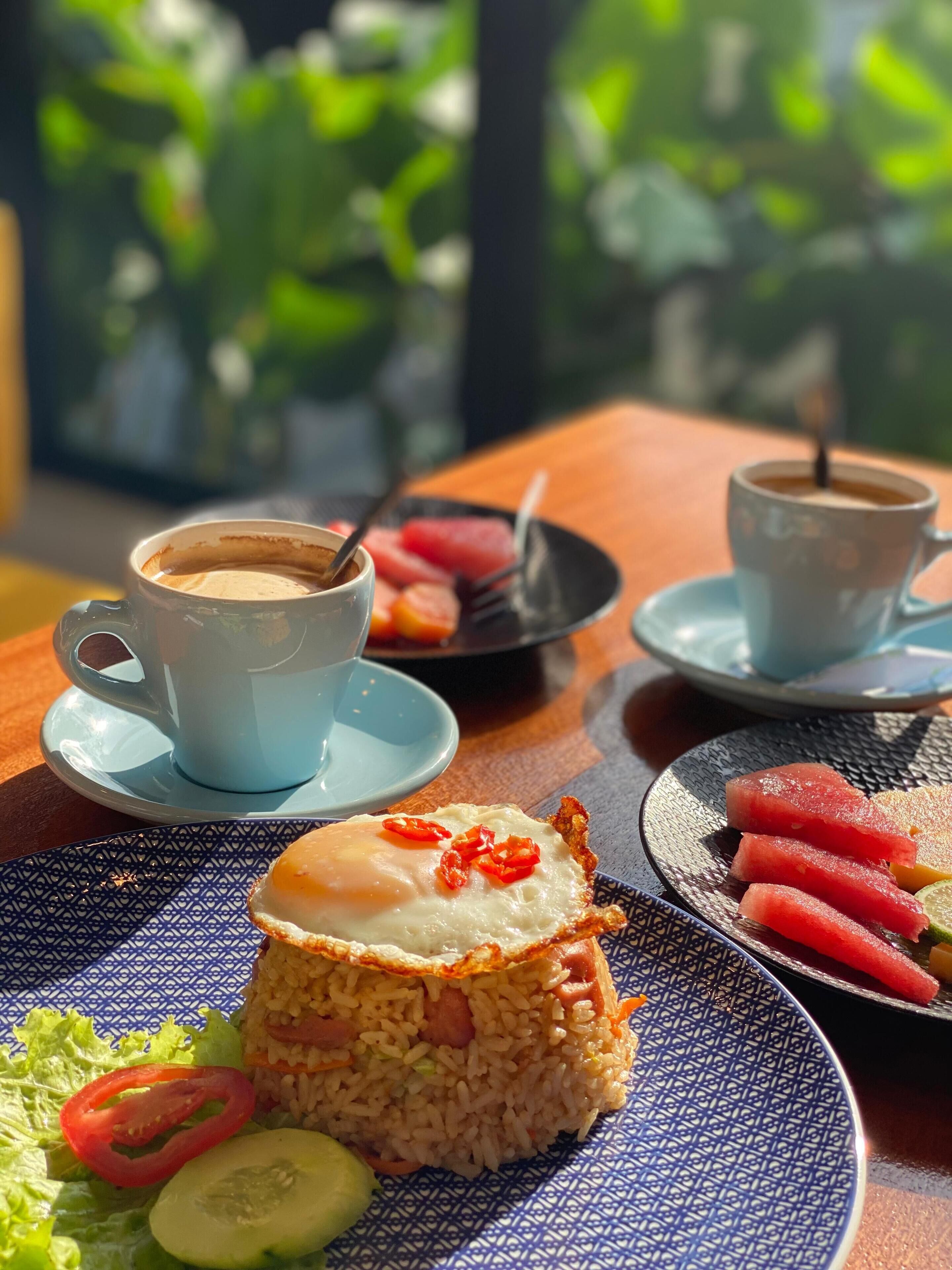A table with a meal consisting of fried rice, a fried egg, and a slice of watermelon, along with two cups of coffee.
