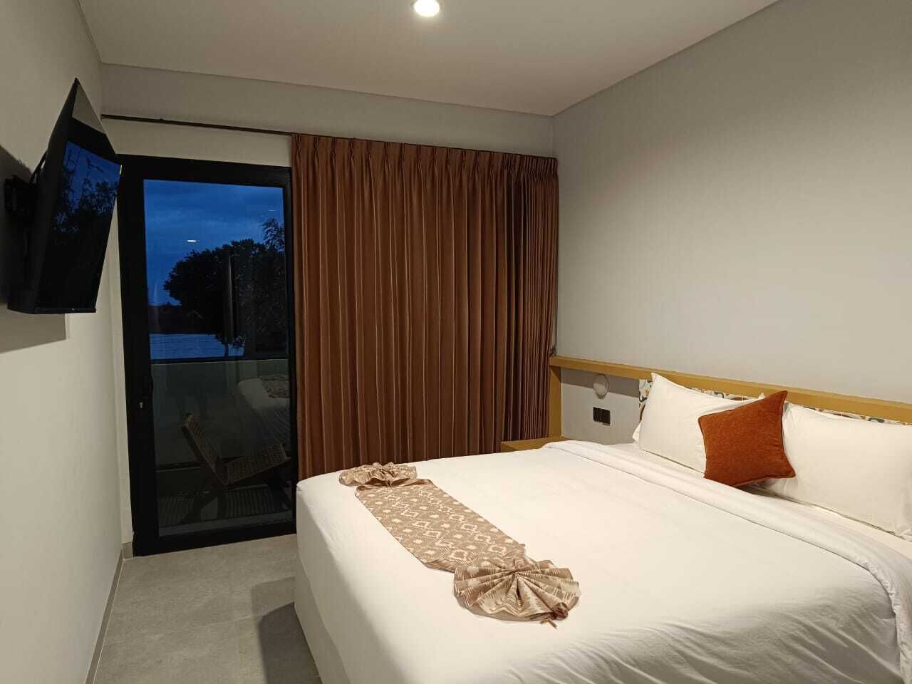 Habitación de hotel con cama, televisor montado en la pared y puerta corrediza de vidrio que da a un espacio exterior.