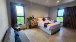 A modern hotel room with a large bed, a sofa, a small table with a plant, and two windows with curtains.