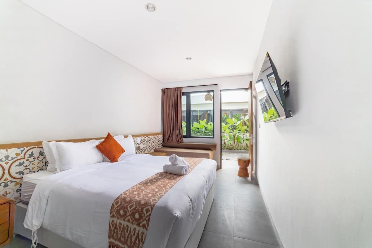 A hotel room with a bed, a TV mounted on the wall, and a window with a view of greenery.