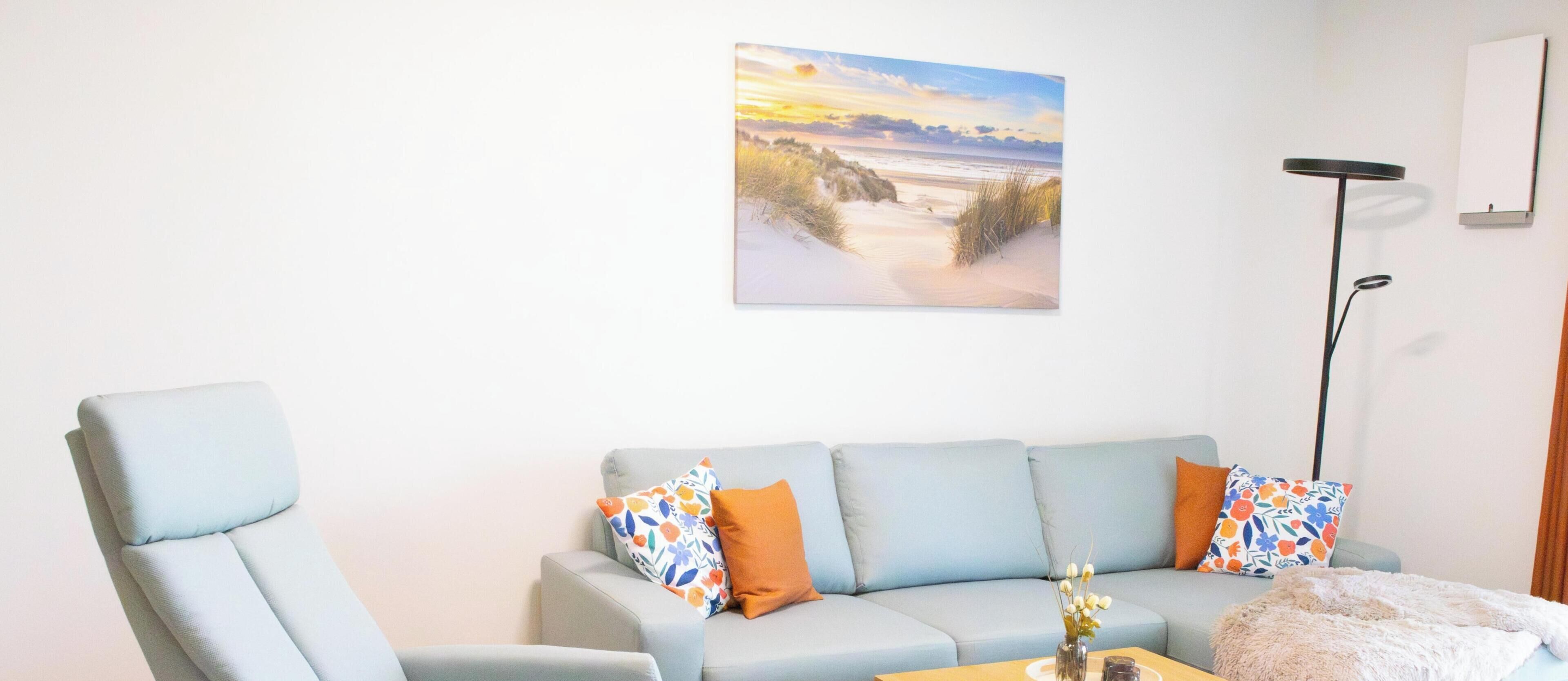 A living room with a light blue sofa, two patterned pillows, a small table with a vase, and a painting of a beach scene on the wall.