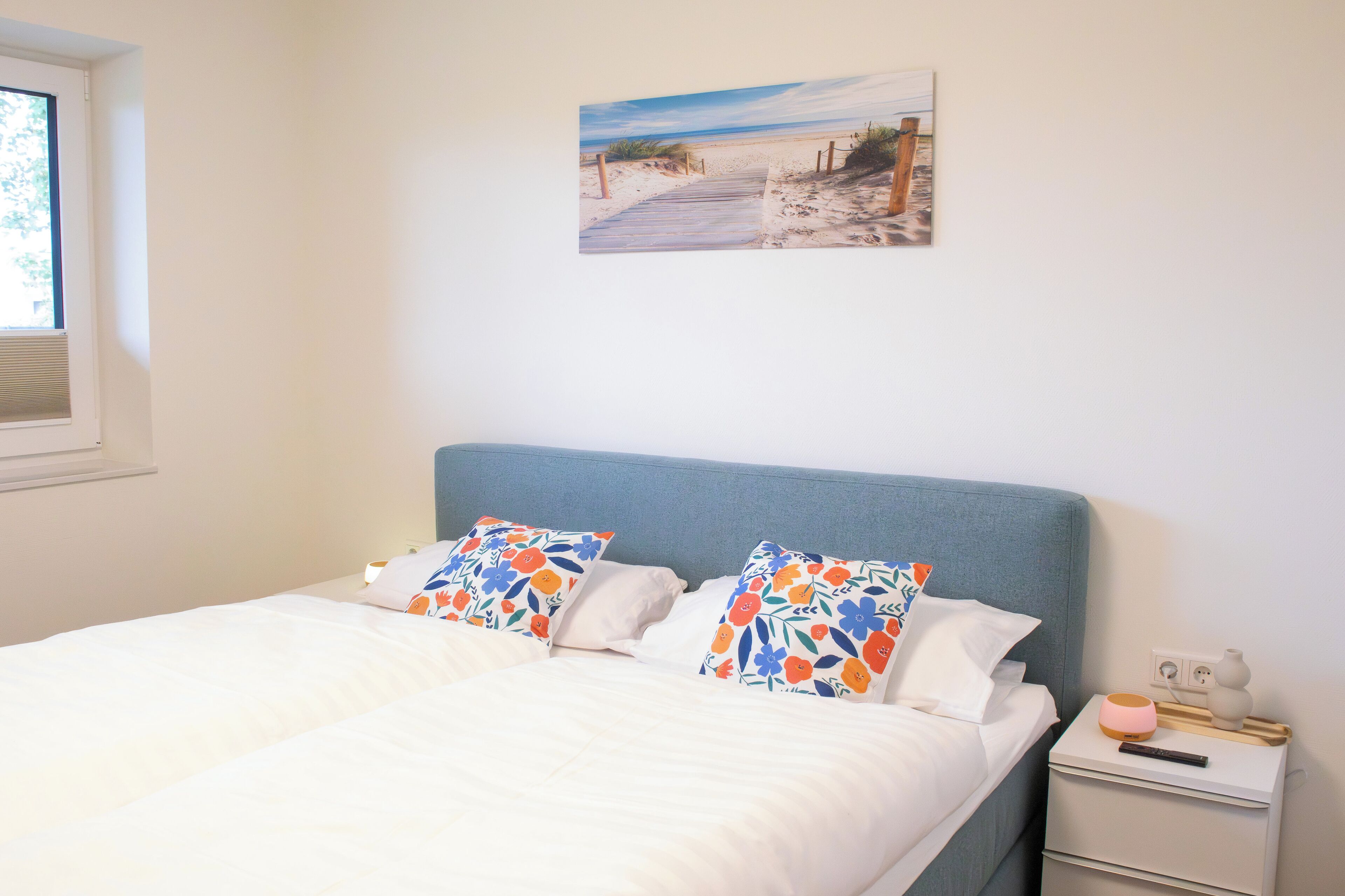 A bedroom with a bed, two decorative pillows, a bedside table with a lamp, and a painting on the wall.