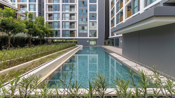A modern building with a swimming pool, surrounded by greenery and featuring balconies with wooden railings. - New Condo Near Beach Prime Location C228 (Choeng Thale)