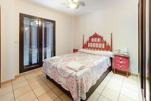 Une chambre à coucher avec un lit, une table de chevet, une tête de lit rouge, une fenêtre avec des stores et un ventilateur de plafond.