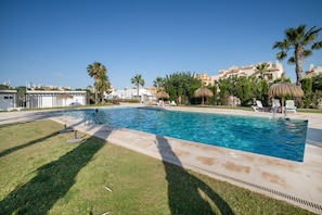 A swimming pool with a clear blue water, surrounded by grass, palm trees, and lounge chairs.