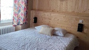 A bedroom with a wooden headboard, a bed with white linens, and a window with floral curtains. - Le commerce d'Ophelie (Corcieux)