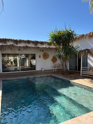 A clear blue pool with a thatched roof structure, a palm tree, and a glass door leading to a poolside area. - Domaine du Douar (Oulad Khallouf)