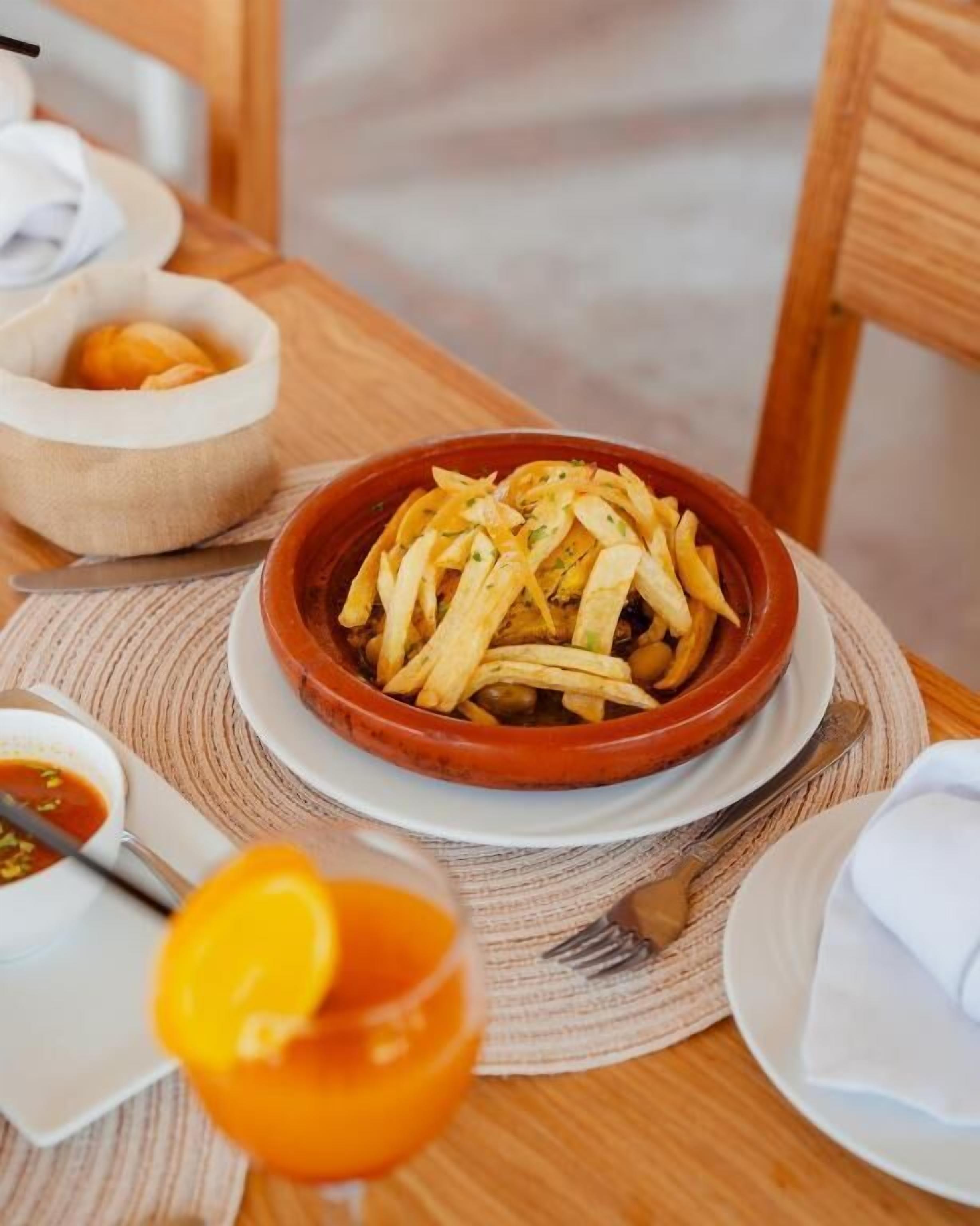 A bowl of fries on a wooden table with a napkin, a spoon, and a glass of orange juice.