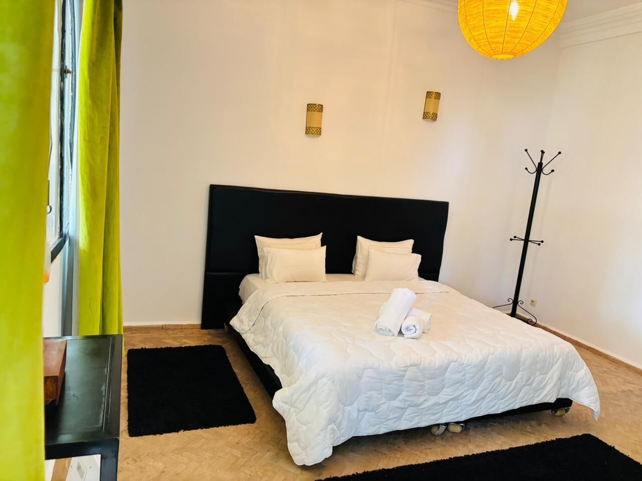 Una cama bien hecha con ropa blanca, cabecera negra y alfombra blanco y negro en el suelo.