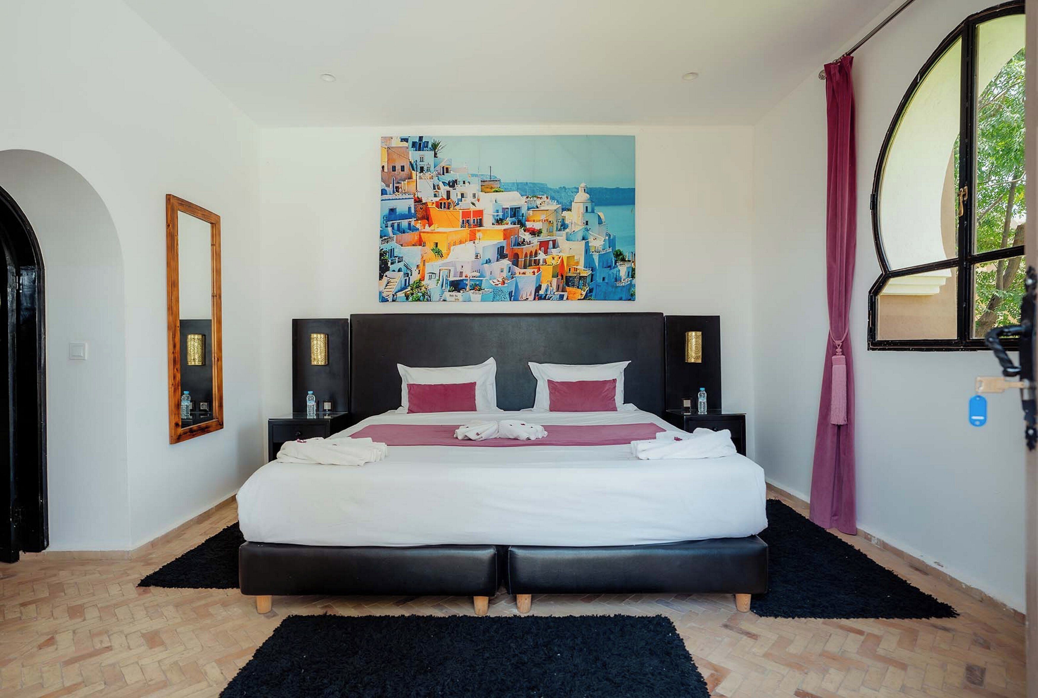 A hotel room with a bed, a painting of a coastal town, a window with curtains, and a door.