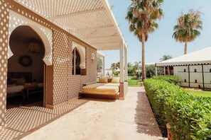A hotel with a covered outdoor seating area, a bed, and a view of palm trees. - Domaine du Douar (Oulad Khallouf)
