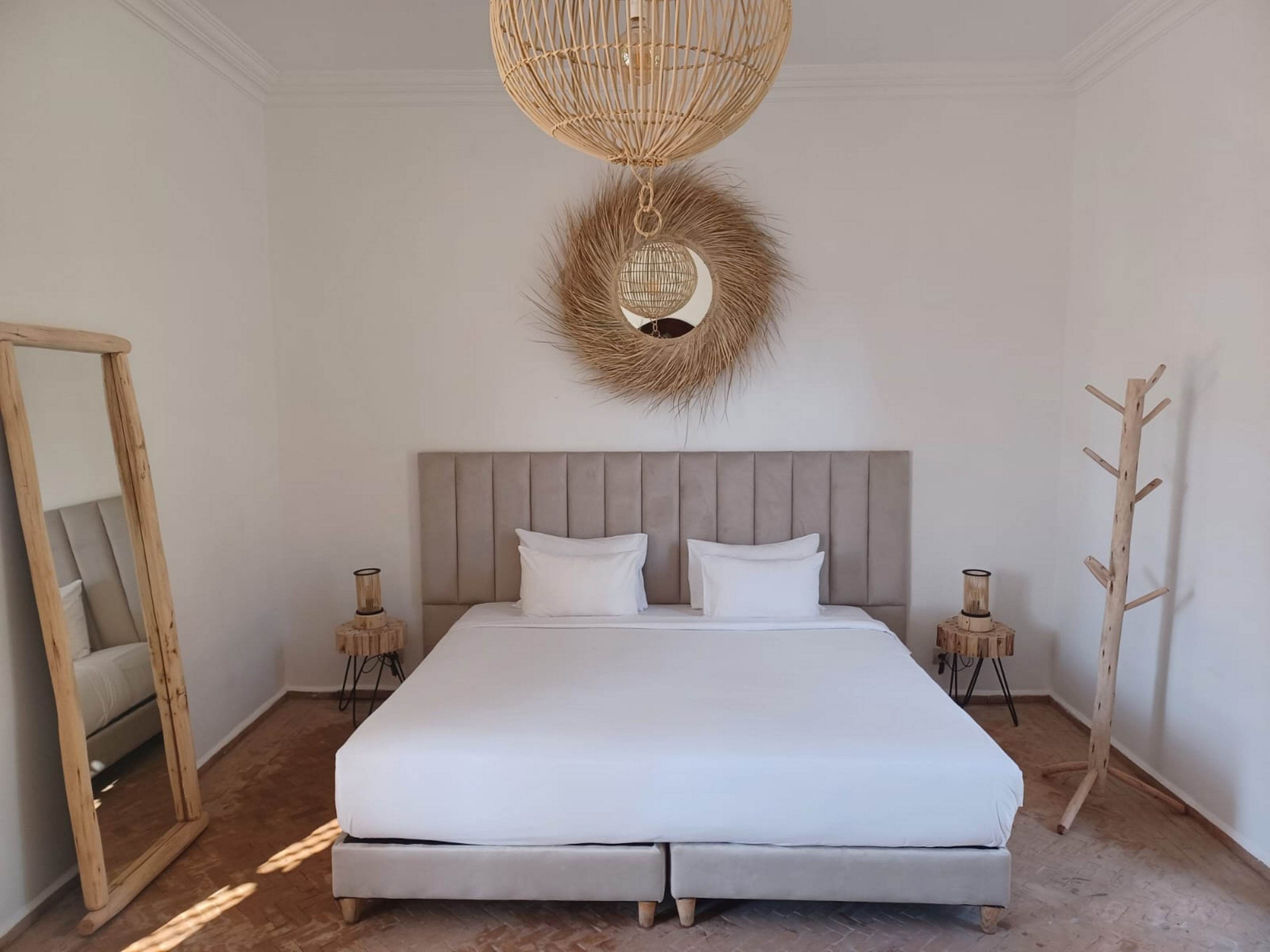 A bedroom with a large bed, a mirror, two bedside tables, a wooden coat rack, and a woven ceiling light fixture.