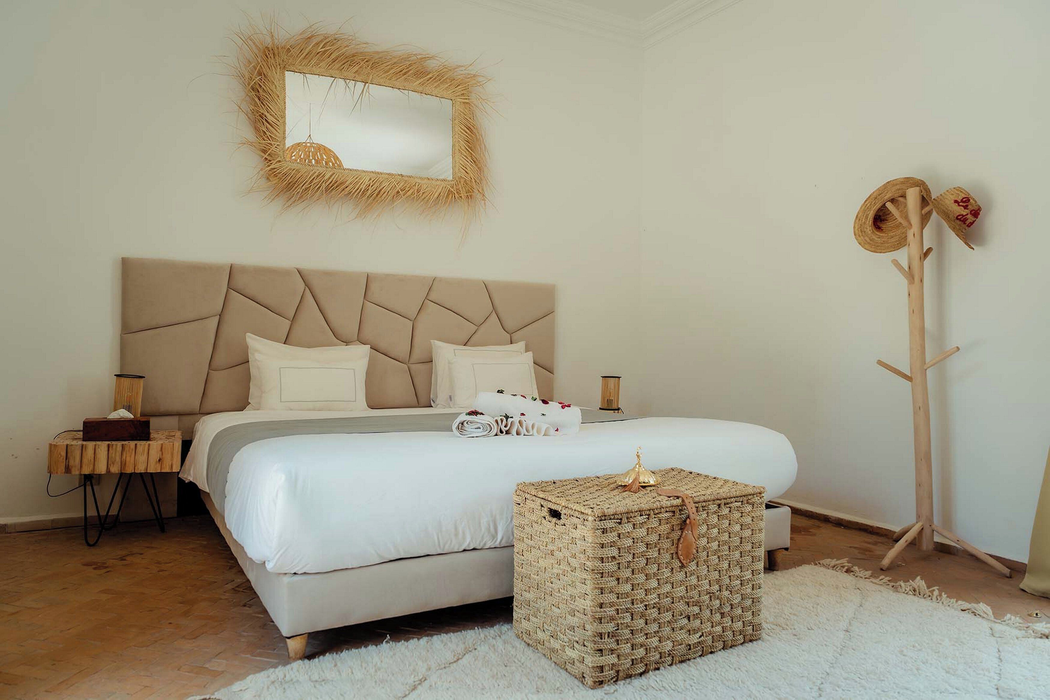 A bedroom with a large bed, a bedside table, a nightstand, a woven ottoman, a mirror with a natural frame, and a wooden coat rack.