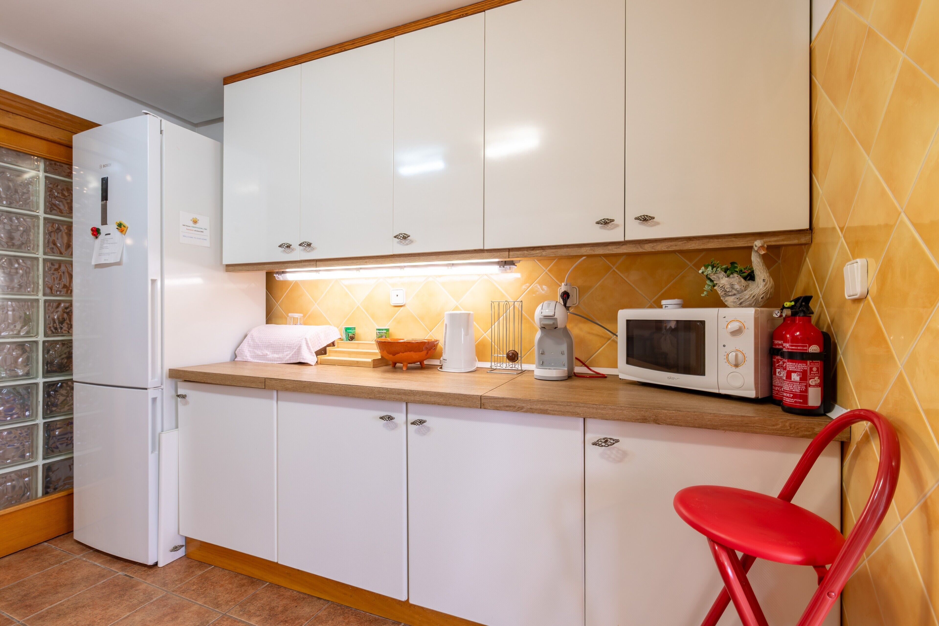 Una cocina con armarios blancos, microondas, tostadora, una silla roja y un extintor.