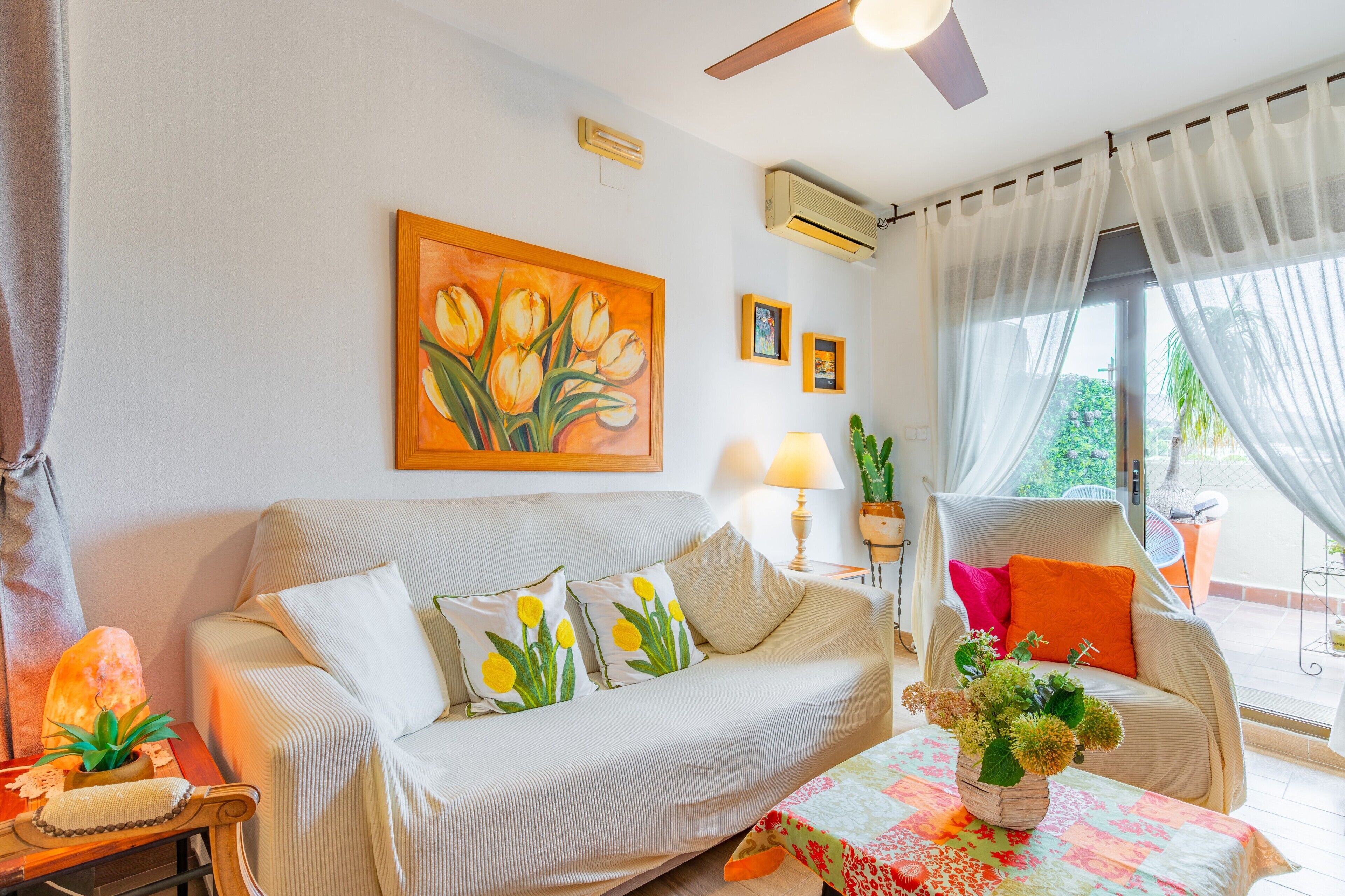 A living room with a sofa, a table with a vase, a painting, and a chair with cushions.