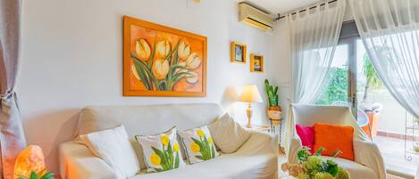 A living room with a sofa, a table with a vase, a painting, and a chair with cushions.