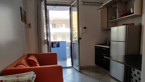 A compact living space with a sofa, a small table, a kitchen area with a refrigerator, and a sliding glass door leading to an outdoor area.