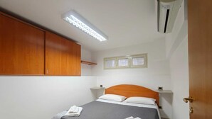 A single bed with a grey bedspread and white pillows, a wooden wardrobe, a small bedside table, and a ceiling light fixture.
