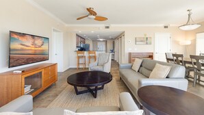 A spacious living room with a large flat-screen TV, a dark wooden coffee table, a beige sofa, and a dining area with a chandelier. - Last-Minute August Beach Deals – Save Big! (Treasure Island)